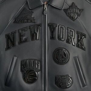 May include: Black leather jacket with the text "NEW YORK" across the chest. The jacket features multiple patches, including "KITH" and basketball-themed designs. A zipper runs down the center.
