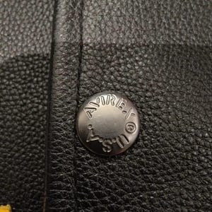 May include: Close-up of a black leather jacket with a silver button. The button is embossed with the word "AMIRI" and "U.S.". Yellow stitching is visible along the bottom edge of the jacket.