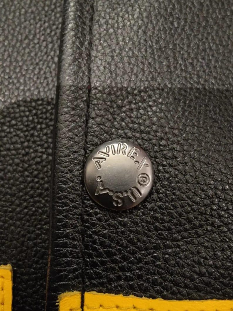 May include: Close-up of a black leather jacket with a silver button. The button is embossed with the word "AMIRI" and "U.S.". Yellow stitching is visible along the bottom edge of the jacket.