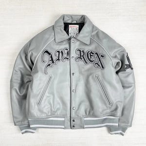 May include: A gray leather jacket with the text "AVI REX" in black script across the chest. The jacket has a collared neckline, button-up front, and white stripes on the cuffs and hem. A small patch is visible on the sleeve.
