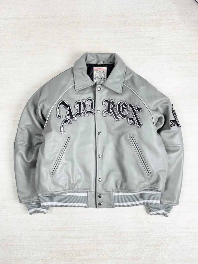 May include: A gray leather jacket with the text "AVI REX" in black script across the chest. The jacket has a collared neckline, button-up front, and white stripes on the cuffs and hem. A small patch is visible on the sleeve.
