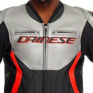 May include: A close-up of a black and silver motorcycle jacket with red accents. The jacket features the brand name "DAINESE" in red lettering across the chest. The jacket has a zippered front and two small chest pockets.