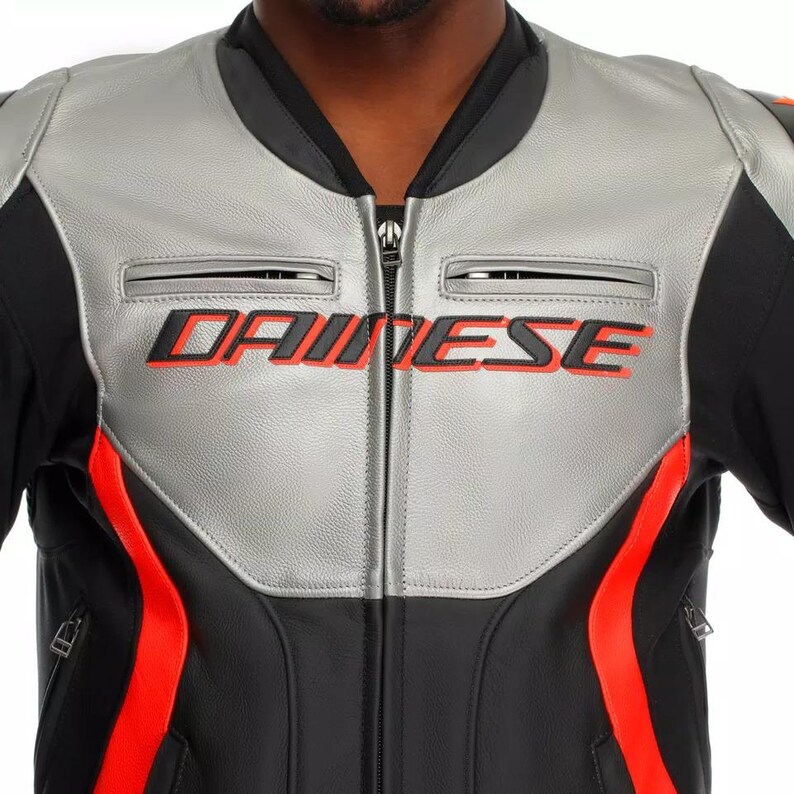 May include: A close-up of a black and silver motorcycle jacket with red accents. The jacket features the brand name "DAINESE" in red lettering across the chest. The jacket has a zippered front and two small chest pockets.