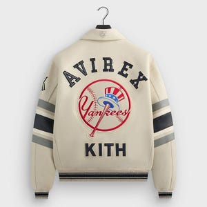 Może przedstawiać: Kremowa kurtka typu varsity z napisem "AVIREX" na górnej części pleców. Duże logo Yankees jest wyśrodkowane poniżej, z napisem "KITH" poniżej. Rękawy mają czarne i szare paski.