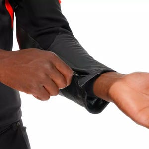 May include: Black jacket with red accents, featuring a zippered sleeve cuff. The jacket appears to be made of a technical fabric, suitable for outdoor activities. The cuff is being unzipped, revealing the wrist.