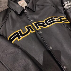 May include: Black leather jacket with a button-up front and a classic collar. The jacket features the word "AURREX" in large yellow and black lettering across the chest. The sleeves have a patch that says "PALACE" and "Equipment".