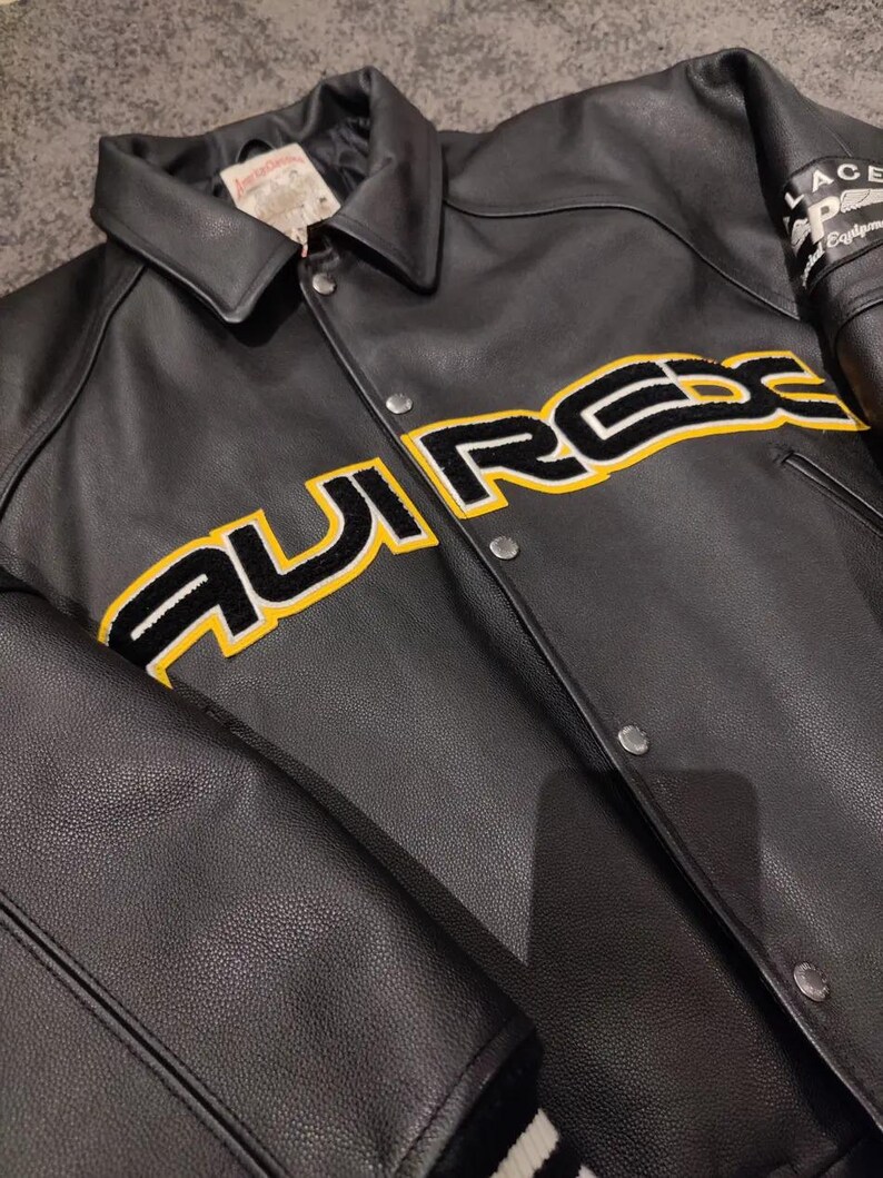 May include: Black leather jacket with a button-up front and a classic collar. The jacket features the word "AURREX" in large yellow and black lettering across the chest. The sleeves have a patch that says "PALACE" and "Equipment".