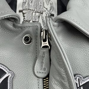 May include: Close-up of a gray leather jacket with a zipper and the brand name "AVIREX" on the zipper pull. The jacket has a "American Classics" label with illustrations of athletes. The jacket is a classic style.
