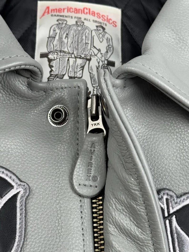May include: Close-up of a gray leather jacket with a zipper and the brand name "AVIREX" on the zipper pull. The jacket has a "American Classics" label with illustrations of athletes. The jacket is a classic style.