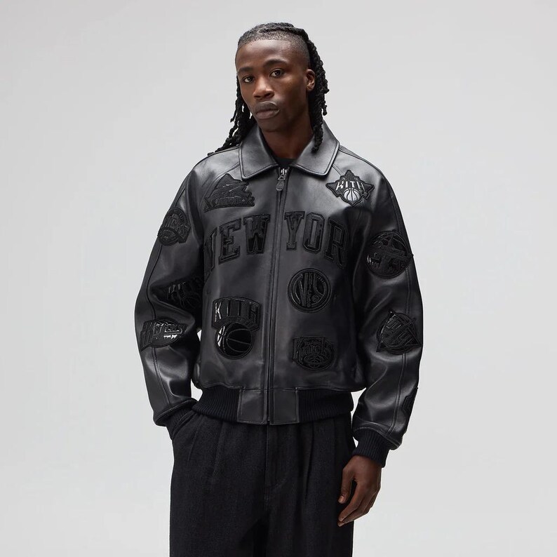 May include: Black leather jacket with multiple patches and the text "NEW YORK" across the chest. The jacket has a zip-up front, a collar, and ribbed cuffs and hem. Patches include basketballs and other sports-related designs.