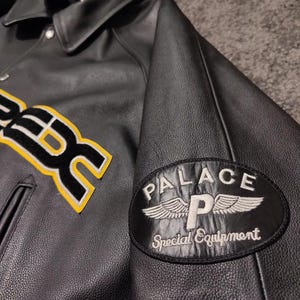May include: Black leather jacket with a button-down collar and a patch on the sleeve that reads "PALACE Special Equipment." The jacket has a yellow and white embroidered design on the front.