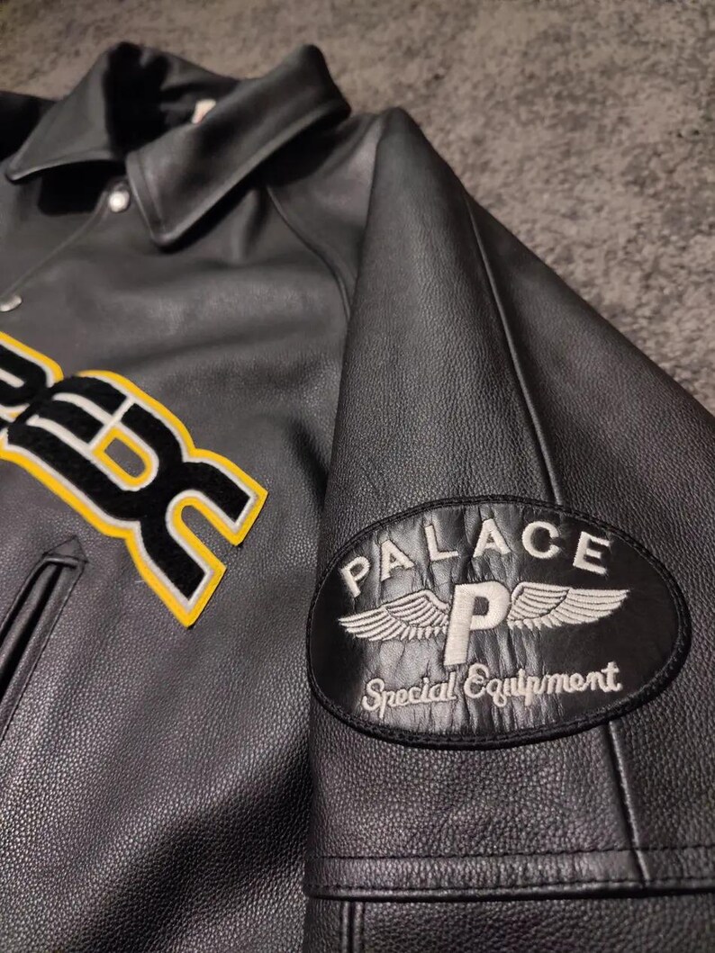 May include: Black leather jacket with a button-down collar and a patch on the sleeve that reads "PALACE Special Equipment." The jacket has a yellow and white embroidered design on the front.