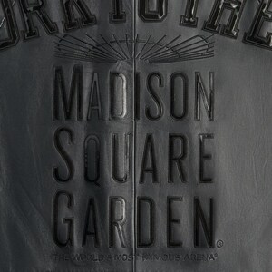 May include: Close-up of a black leather jacket with the embossed text "MADISON SQUARE GARDEN." The text is in a bold, serif font. Below the text is the phrase "THE WORLD'S MOST FAMOUS ARENA." The jacket has a textured surface.