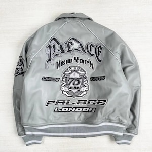 May include: A gray leather jacket with white trim and the text "PALACE New York" in a stylized font. Additional text includes "LONDON", "TOKYO", and "75". The jacket has a ribbed hem and cuffs with white stripes.