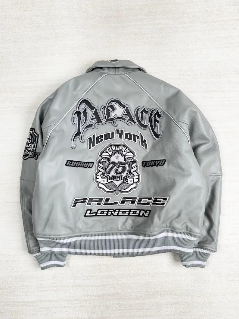 May include: A gray leather jacket with white trim and the text "PALACE New York" in a stylized font. Additional text includes "LONDON", "TOKYO", and "75". The jacket has a ribbed hem and cuffs with white stripes.