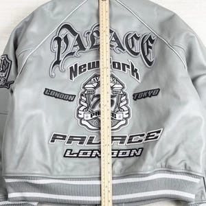 May include: A light gray bomber jacket with white and black accents. The jacket features the word "PALACE" in a gothic font, along with "New York", "London", and "Tokyo". The jacket has a striped hem and cuffs.