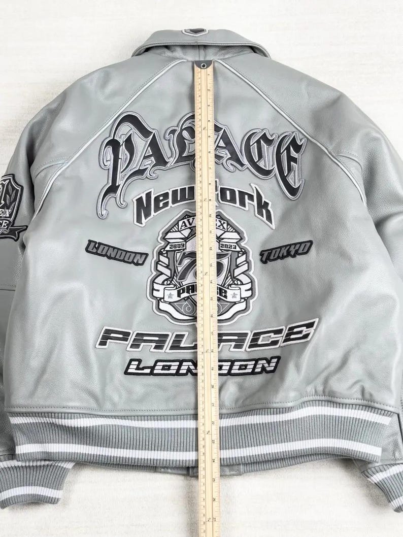May include: A light gray bomber jacket with white and black accents. The jacket features the word "PALACE" in a gothic font, along with "New York", "London", and "Tokyo". The jacket has a striped hem and cuffs.