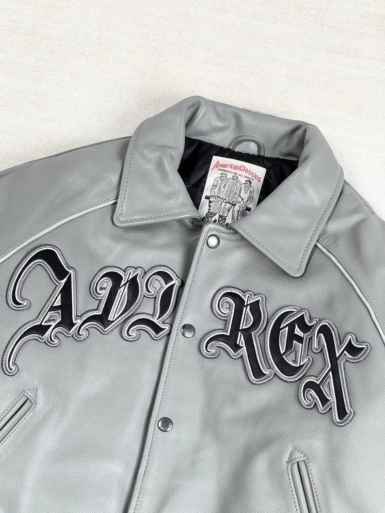 May include: A gray leather jacket with the text "AVL REX" in black script. The jacket has a collar, snap closures, and white trim. The label reads "American Classics."