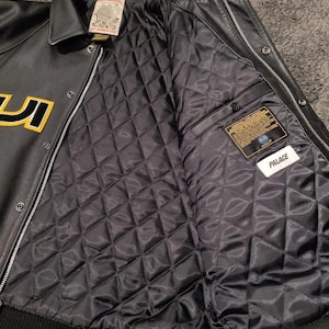May include: Black leather jacket with a quilted black lining. The jacket features a button-up front, a collar, and a ribbed hem. The jacket has yellow lettering and a "Palace" tag.