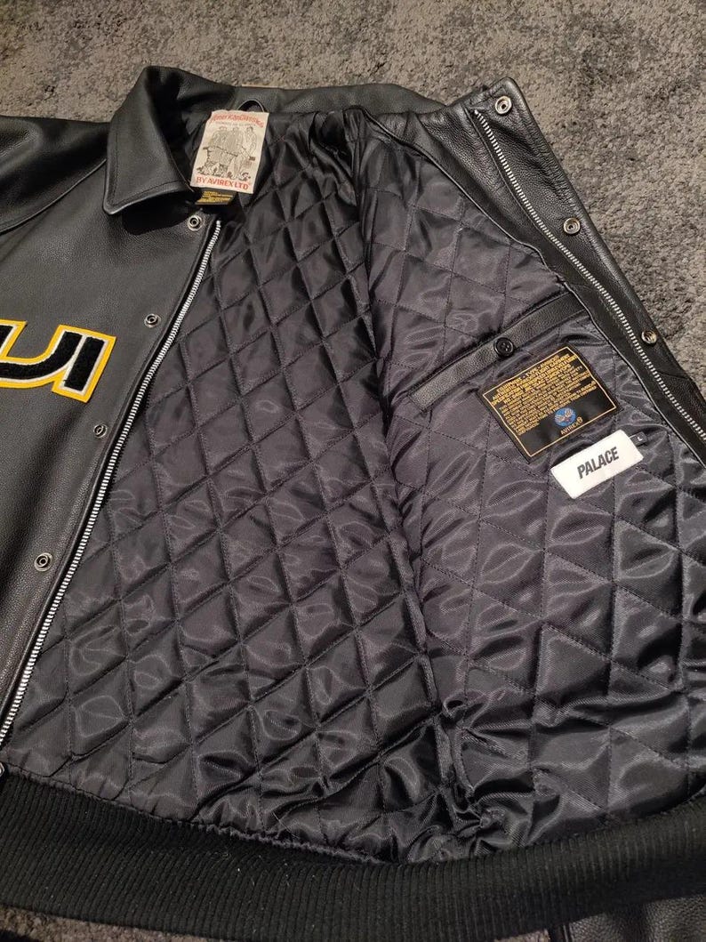 May include: Black leather jacket with a quilted black lining. The jacket features a button-up front, a collar, and a ribbed hem. The jacket has yellow lettering and a "Palace" tag.