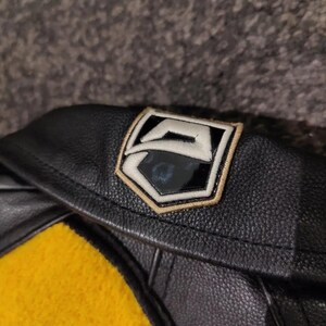 May include: Black leather jacket with a yellow felt detail. The jacket has a patch with a white and black design, edged in gold. The image shows the collar and part of the jacket's body.