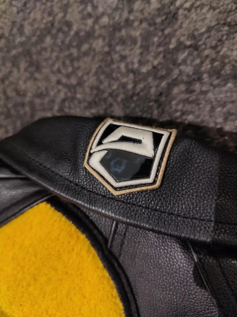 May include: Black leather jacket with a yellow felt detail. The jacket has a patch with a white and black design, edged in gold. The image shows the collar and part of the jacket's body.