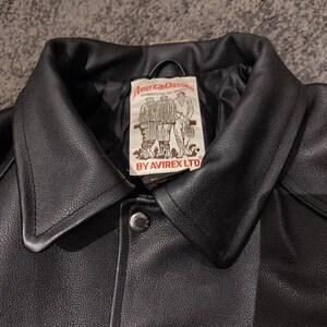 May include: A close-up of a black leather jacket with a wide collar and a button closure. The jacket has a label with the text "American Classics" and illustrations of people playing sports. The brand name "AVIREX LTD" is also visible.