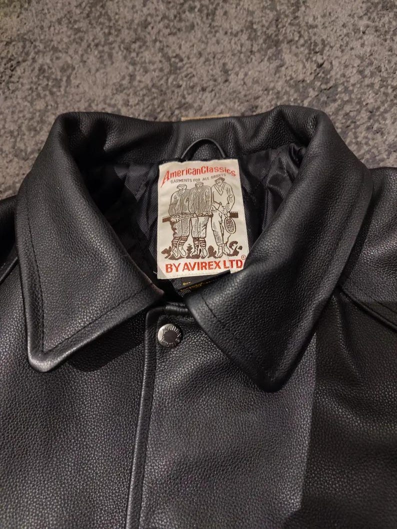 May include: A close-up of a black leather jacket with a wide collar and a button closure. The jacket has a label with the text "American Classics" and illustrations of people playing sports. The brand name "AVIREX LTD" is also visible.