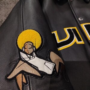 May include: Black leather jacket with a cartoon patch of a figure with a yellow afro and white coat. The jacket has a button-up front and a collar. The letters "U" and "S" are visible in yellow and black.