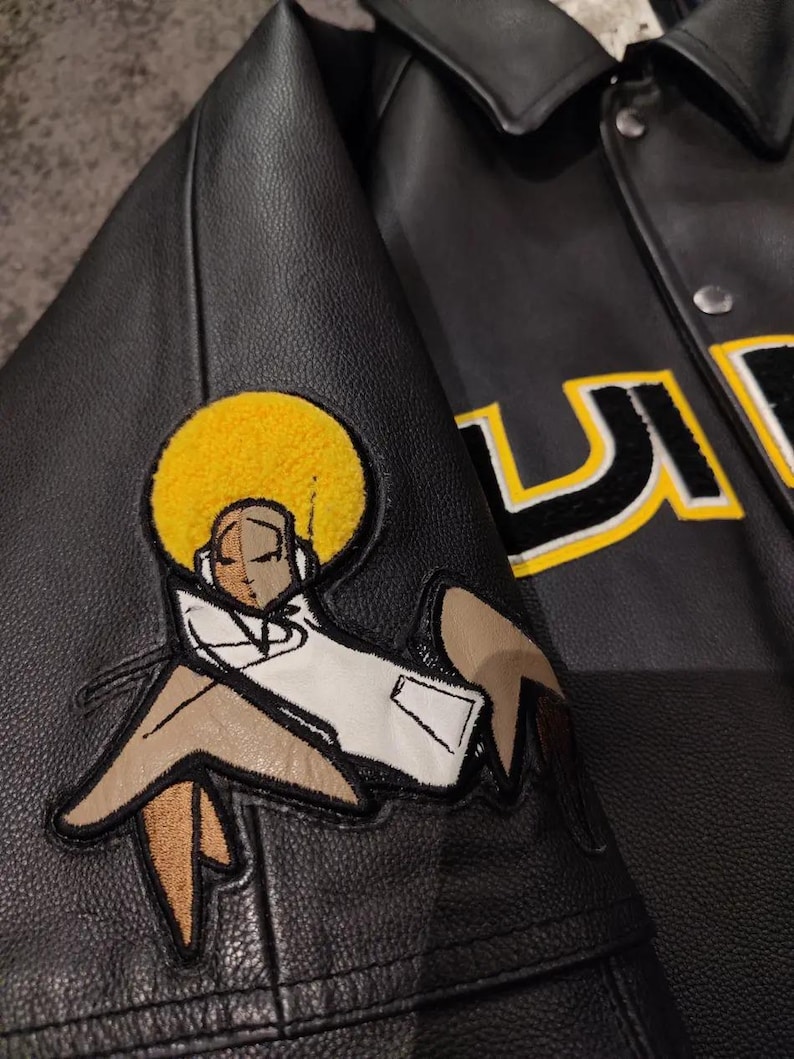 May include: Black leather jacket with a cartoon patch of a figure with a yellow afro and white coat. The jacket has a button-up front and a collar. The letters "U" and "S" are visible in yellow and black.