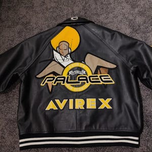 May include: Black leather jacket with a large graphic on the back. The graphic includes a stylized figure, the word "PALACE" in yellow, and the "AVIREX" logo. The jacket has a striped hem.
