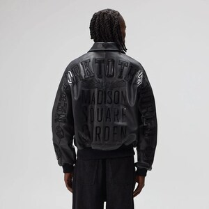 May include: Black leather bomber jacket with embossed text on the back that reads "NEW YORK TO THE MADISON SQUARE GARDEN". The jacket has a ribbed hem and cuffs. The model is wearing black pants.