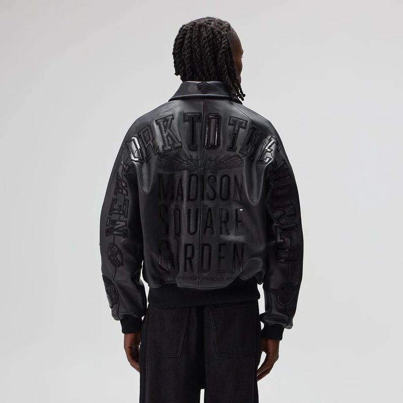 May include: Black leather bomber jacket with embossed text on the back that reads "NEW YORK TO THE MADISON SQUARE GARDEN". The jacket has a ribbed hem and cuffs. The model is wearing black pants.