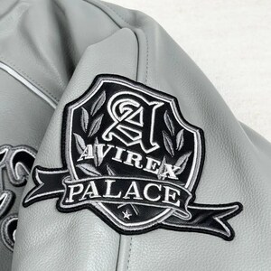 May include: Close-up of a gray leather jacket with a black and white embroidered patch. The patch features the words "AVIREX PALACE" in white lettering, with a stylized emblem and a star.