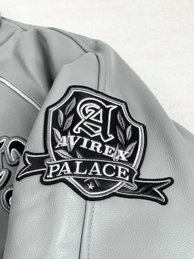 May include: Close-up of a gray leather jacket with a black and white embroidered patch. The patch features the words "AVIREX PALACE" in white lettering, with a stylized emblem and a star.