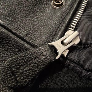 May include: Close-up of a black leather jacket showing a silver zipper and snap closure. The zipper pull is textured and the zipper teeth are visible. The leather has a pebbled texture.