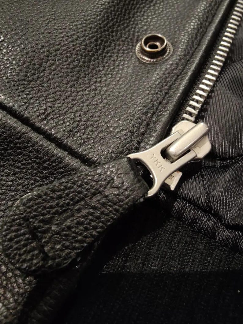 May include: Close-up of a black leather jacket showing a silver zipper and snap closure. The zipper pull is textured and the zipper teeth are visible. The leather has a pebbled texture.