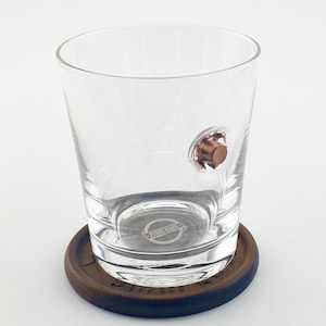 May include: A clear glass tumbler with a copper-colored bullet embedded in the side, resting on a dark brown wooden coaster. The coaster has a dark blue edge and a logo on the bottom of the glass.