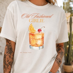 Pode incluir: Uma t-shirt branca com o texto laranja "Old Fashioned GIRLIE" acima de um gráfico de um cocktail Old Fashioned num copo com gelo, uma fatia de laranja e uma cereja, com o texto "est. happy hour" impresso abaixo do copo.