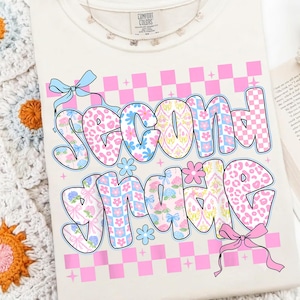 Può includere: Una t-shirt color crema con la scritta Second Grade in grandi lettere stile bolla. Le lettere sono riempite con stampa leopardata rosa, motivi a scacchi blu e rosa e disegni floreali. Sono presenti un fiocco azzurro e un fiocco rosa.