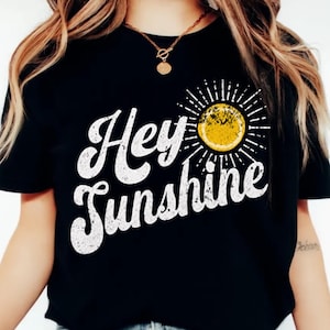 Puede incluir: Camiseta negra con la frase "Hey Sunshine" en letras blancas desgastadas. Un sol amarillo con rayos blancos está a la derecha del texto. La camiseta está hecha de un material suave.