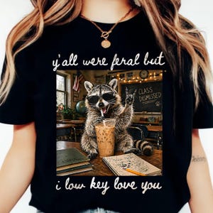 Puede incluir: Camiseta negra con un diseño de un mapache con gafas de sol, bebiendo de un vaso y haciendo el gesto de rock. El texto en la camiseta dice "y'all were feral but i low key love you".