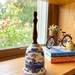 May include: A ceramic bell with a dark wooden handle and a blue and white Dutch windmill design. The bell is displayed on a wooden shelf with other decorative items, including a miniature bicycle and books.