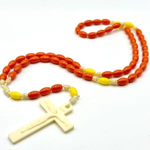 May include: Orange and yellow beaded rosary with a cream-colored crucifix. The rosary features oval-shaped beads in orange and yellow, strung together with a cream-colored cord. The crucifix depicts a figure and is made of a cream-colored material.