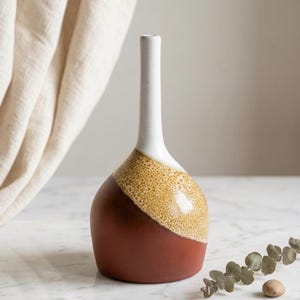 May include: A ceramic vase with a white neck, a textured tan band, and a reddish-brown base. The vase has a rounded body and a long, slender neck. The background includes a white curtain and a sprig of eucalyptus.