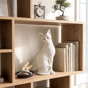 May include: A white ceramic cat figurine sits on a light oak shelf next to a stack of books with titles including Wabi-Sabi Welcome and Ceramic Form. A small black bowl containing smooth gray stones and dried grass stems sits beside the cat.
