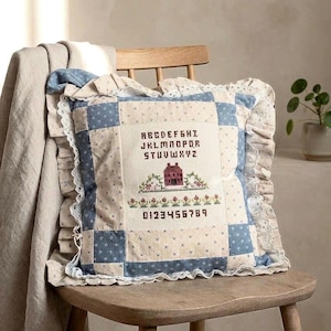 May include: A square throw pillow featuring a patchwork design in blue and cream floral patterns. The center panel displays a cross-stitched alphabet, numbers 0-9, and a red house motif. The pillow is finished with a ruffled edge and white lace trim.