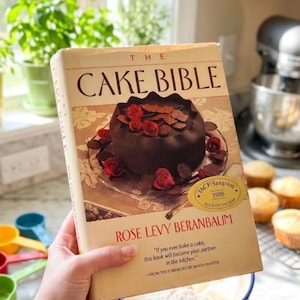 May include: A white ceramic mug with a black handle and the text "But First, Coffee" in a bold, black font. The mug is set against a blurred background, suggesting a cozy setting.