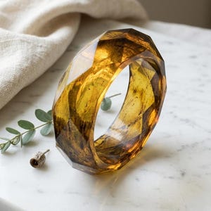 May include: A faceted, amber-colored resin bangle bracelet. The bracelet has a geometric design with multiple flat surfaces. The transparent material allows light to pass through, revealing internal patterns. The bracelet is displayed on a white marble surface.