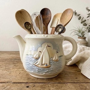 May include: A cream-colored ceramic teapot-shaped utensil holder with a nautical design of sailboats on the sea. The holder is filled with wooden spoons, a whisk, and measuring spoons. The teapot has a handle and spout.